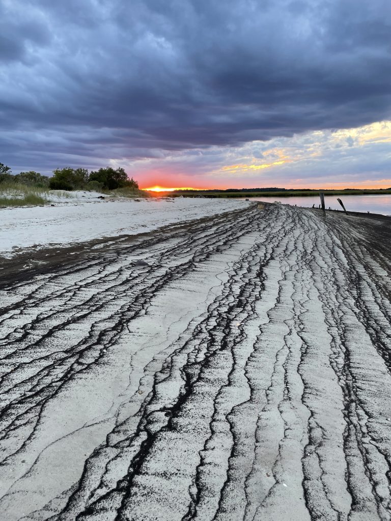 black tide lines on pale sand stretch towards a sliver of red sunset under dark approaching clouds.